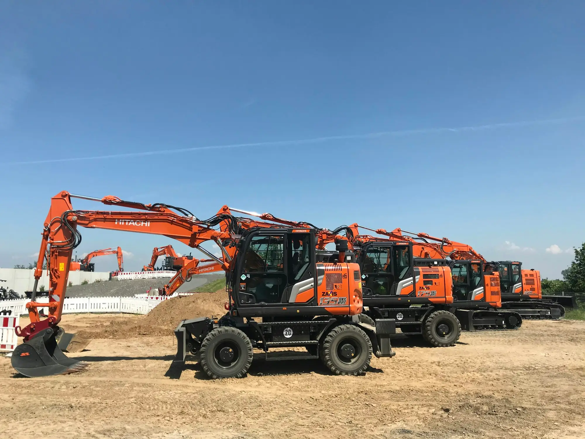 Row of new wheeled excavators lined up on a worksite under a clear blue sky